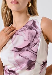 Top bianco senza maniche con motivo floreale in rosa e viola, caratterizzato da una texture satinata liscia e scollatura rotonda. Orecchini a cerchio dorati visibili.
