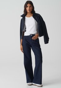 Jeans in denim blu scuro a gamba larga abbinati a una maglietta bianca aderente e una giacca con zip blu navy, presentati in una posa casual.