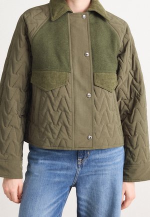 Persoon draagt een olijfgroene doorgestikte jas met drukknopen, ribfluwelen accenten, voorzakken en blauwe spijkerbroek zichtbaar vanaf de taille naar beneden.