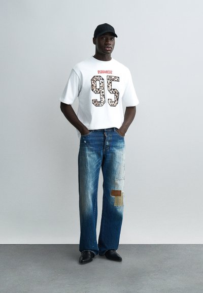 Άνδρας που φοράει λευκό υπερμεγέθες T-shirt με λεοπάρ στάμπα "95" και μπλε τζιν με patches, στέκεται με τα χέρια στις τσέπες μπροστά σε απλό φόντο.