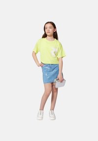 Camiseta corta de color amarillo neón con logo blanco, falda de mezclilla azul claro con lentejuelas, bolso plateado y zapatos de plataforma blancos.