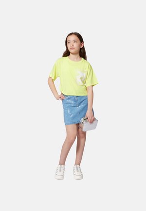 Camiseta corta de color amarillo neón con logo blanco, falda de mezclilla azul claro con lentejuelas, bolso plateado y zapatos de plataforma blancos.