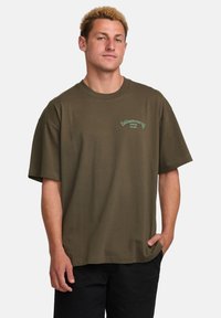 Olive green cotton T-shirt with short sleeves, crew neckline, and small white logo on the left chest. Casual fit, smooth texture.