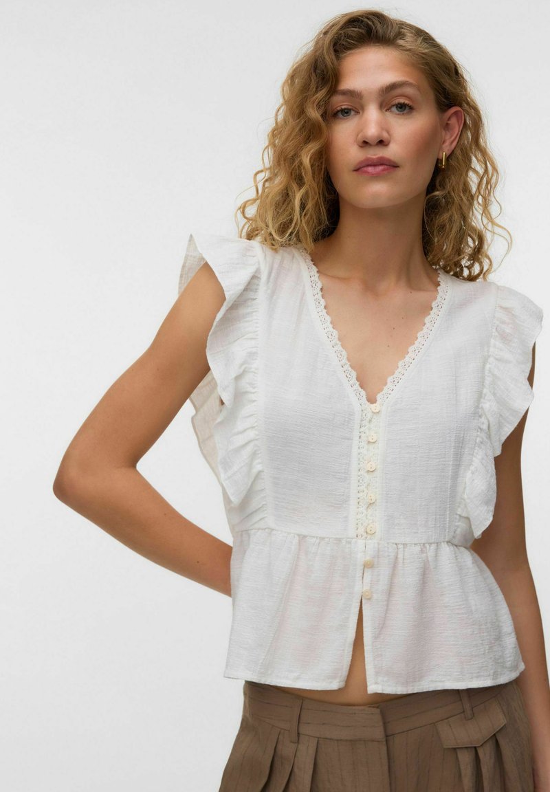 Femme aux cheveux bouclés portant un chemisier blanc sans manches à volants avec des boutons et un pantalon plissé beige sur un fond uni.