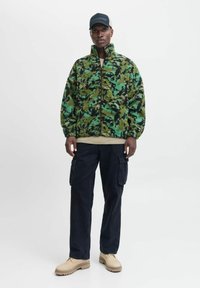 Fleecejack in groen en zwart camo, met een ritssluiting aan de voorkant en een hoge kraag. Gecombineerd met zwarte cargobroek en crèmekleurige schoenen.