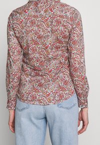 Femme portant une chemise ajustée à manches longues avec des motifs cachemire rouges et roses, associée à un jean en denim bleu clair, vue de dos.