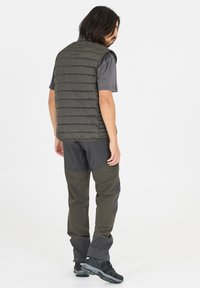 Grön pufferväst med vertikala sömmar, bärs över en grå t-shirt med kort ärm och kombineras med mörkgrå byxor och svarta vandringsskor.