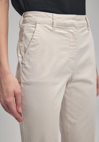 Pantalons en coton beige avec une texture lisse, présentant des poches latérales et une coupe décontractée. La taille est structurée avec des accents de couture visibles.
