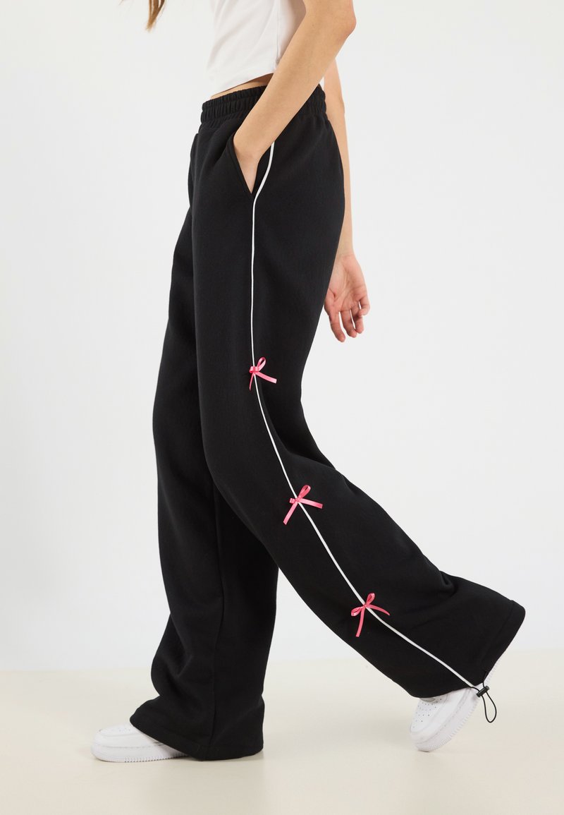 Black sweatpants with white piping detail and pink bows along the side, featuring a relaxed fit and side pockets. Paired with white sneakers.