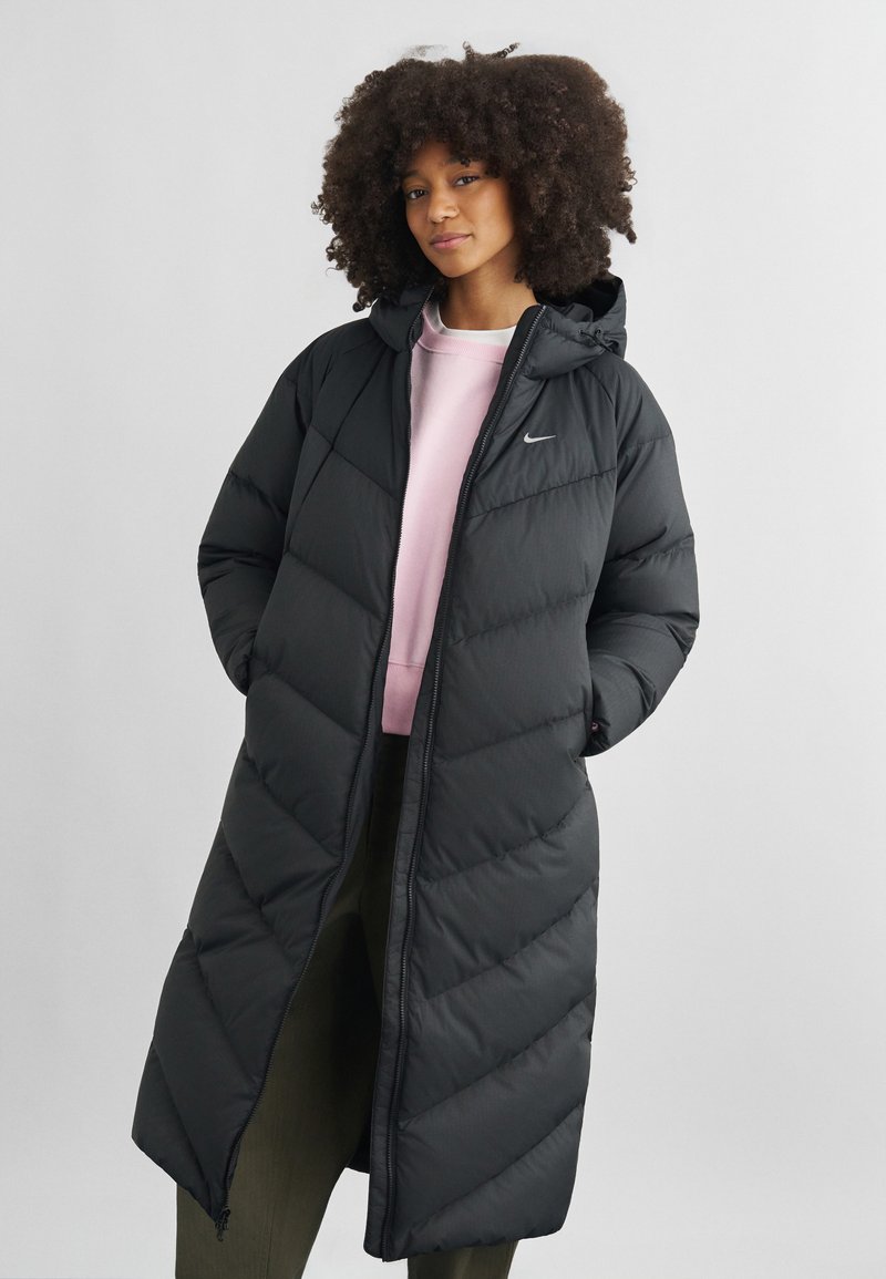 Frau mit lockigem Haar, die eine lange schwarze gesteppte Jacke über einem rosa Sweatshirt und dunklen Hosen trägt, mit den Händen in den Taschen stehend.