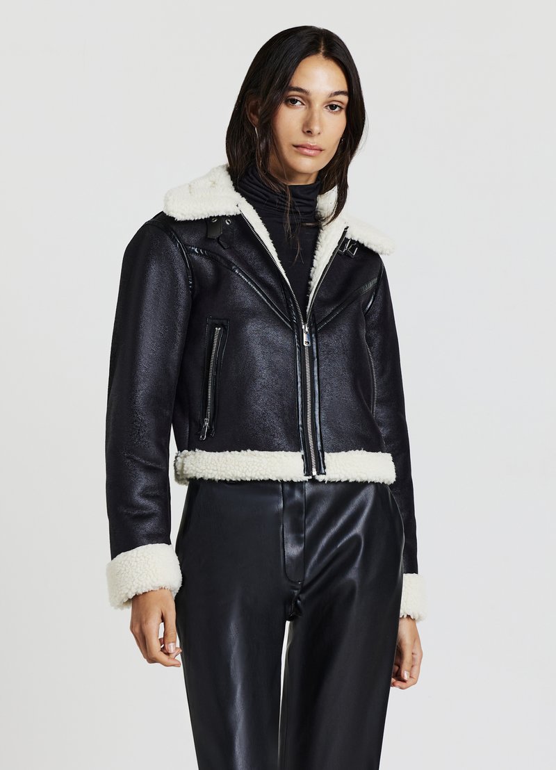 Veste en cuir noire courte avec un col et des poignets en peau de mouton, dotée d'une fermeture éclair et de poches latérales. La texture est lisse et brillante.