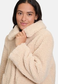 Płaszcz z polarowego materiału w jasnym beżu, z szerokim kołnierzem i luźnym krojem, o teksturowanej, puszystej powierzchni i szerokich rękawach.