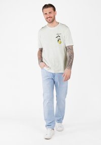 Glimlachende man met tatoeages op beide armen draagt een lichtgrijze T-shirt met citroenprint, lichtblauwe jeans en witte sneakers, staande op een witte achtergrond.