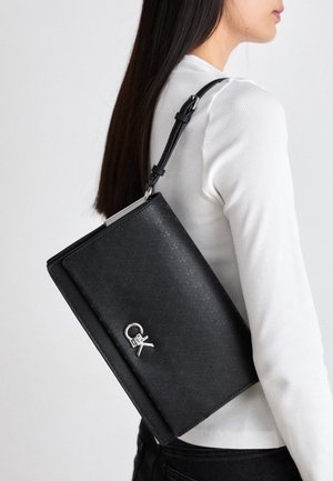 Vrouw met lang donker haar, gekleed in een wit geribd topje en zwarte broek, draagt een zwarte gestructureerde schoudertas met een zilveren gesp.