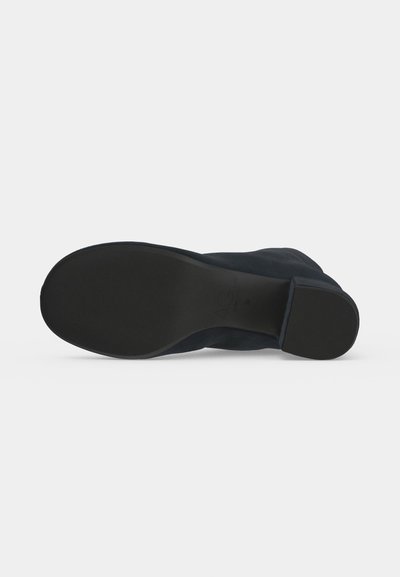Semelle de chaussure noire avec un petit talon carré, vue par dessous sur un fond clair uni.