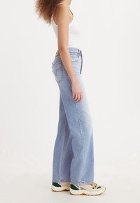 Light blue wide-leg jeans made of denim, featuring a high waist, four pockets, and a slight faded texture. Paired with neutral athletic shoes.