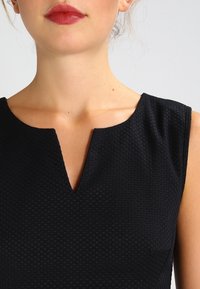 Robe noire avec un motif texturé en nid d'abeille ; présente un décolleté en V et un design sans manches. Le tissu semble lisse avec un léger éclat.