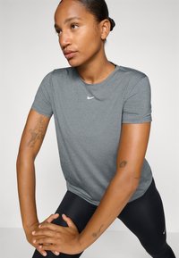 Camisa atlética corta de manga gris con cuello redondo y un pequeño logo de Nike blanco en el pecho. El modelo lleva leggings negros.