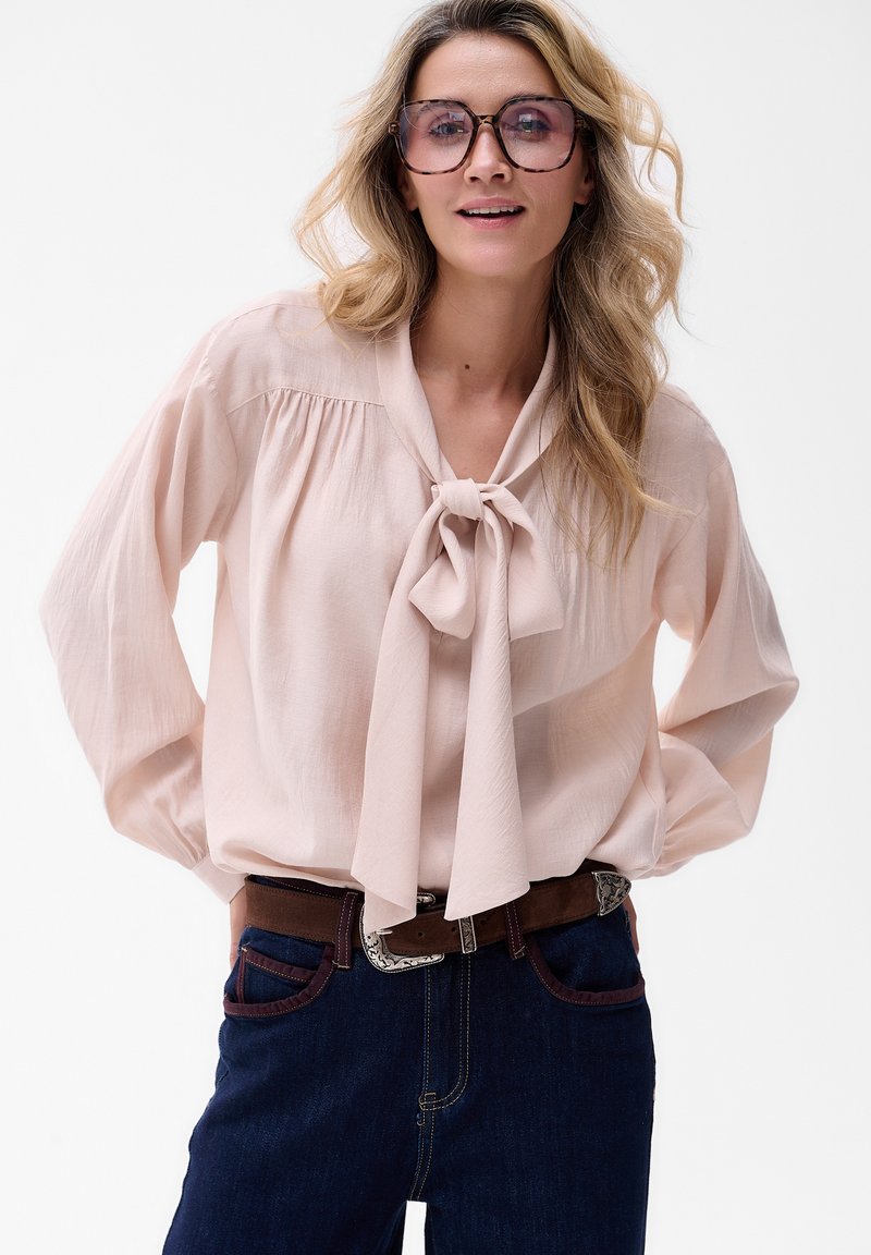 Jasnoróżowa bluzka z detalem w postaci kokardy, długie rękawy i marszczone ramiona, zestawiona z ciemnoniebieskimi dżinsami oraz brązowym paskiem z srebrnymi elementami.