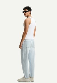 Top alb, blugi deschiși, lejerați, cu buzunare pe spate și adidași crem; modelul este întors lateral, purtând ochelari de soare negri.
