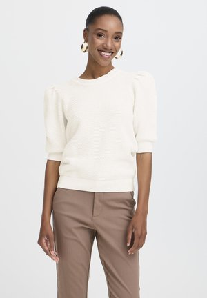 Suéter blanco texturizado con mangas abullonadas, escote redondo y dobladillo acanalado, combinado con pantalones marrones de corte ajustado. Pendientes de aro dorados.