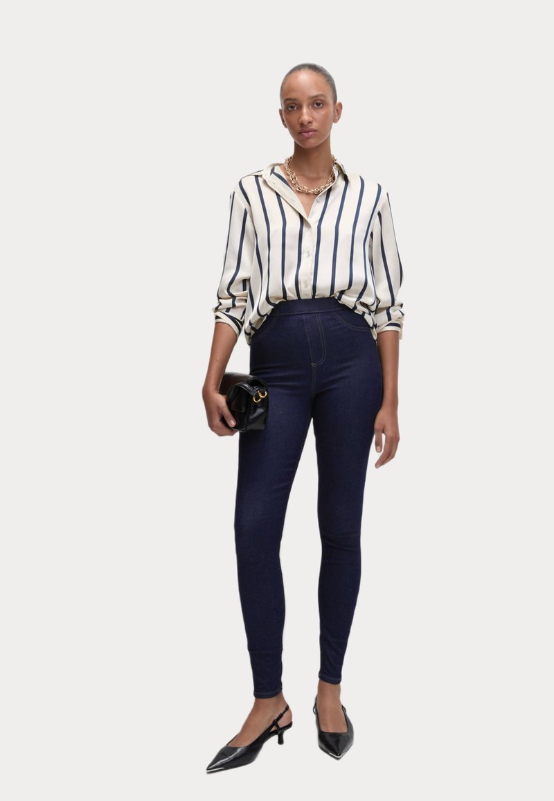 Randig, knappad skjorta i krämfärgad och marinblå; högmidjade mörka denimleggings; svart kuvertväska; spetsiga svarta klackar. Slät tyg och figursydd design.