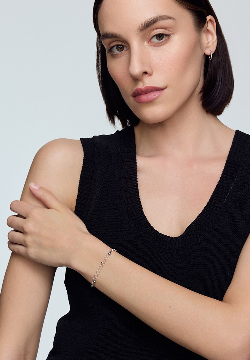 Srebrna bransoletka z delikatnym łańcuchem, z małymi akcentami i zapięciem, noszona na nagim nadgarstku w zestawieniu z czarnym swetem.