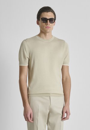 Giovane uomo con occhiali da sole neri, maglione beige a maniche corte e pantaloni beige coordinati, su uno sfondo chiaro e uniforme.