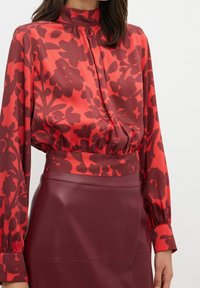 Rød blomsterbluse med høj hals, plisseringer og lange ærmer, parret med en bordeaux læderskirt. Glat tekstur og livlig farvekontrast.