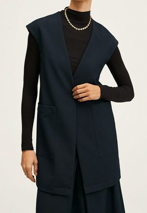 Personne portant un gilet sans manches bleu marine avec deux poches avant sur un haut noir à manches longues et un collier chaîne doré.
