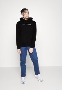 Svart hettegenser med "HILFIGER"-logo, kombinert med blå bukser. Skoene er hvite med flerfargede detaljer. Enkle, avslappede antrekk.