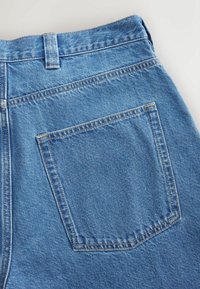 Zone de la poche arrière et de la ceinture d'un jean en denim bleu avec des coutures visibles sur un fond clair.