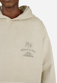Sweatshirt capucho bege com um acabamento texturizado. O gráfico frontal inclui o logótipo "PDR" e "Sussurros do Frio" com um design de montanha e sol.