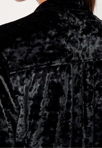 Camicia in velluto nero con una superficie morbida e testurizzata, caratterizzata da un motivo sottile e un design a collare classico. In evidenza cuciture strutturate.