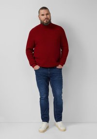 Czerwony, prążkowany sweter z golfem zestawiony z ciemnoniebieskimi dżinsami i białymi trampkami. Osoba stoi na tle jednolitego tła.