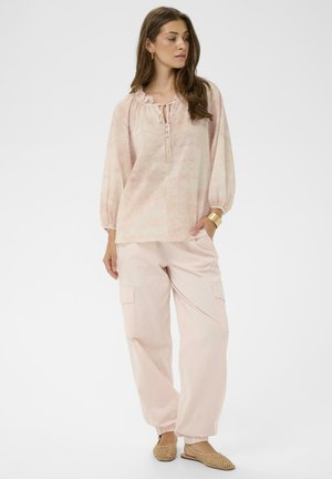 Fiatal nő laza rózsaszín blúzban és zsebes nadrágban, bézs színű szőtt lapos cipőben, egyik kezét zsebre téve, fehér háttér előtt áll.