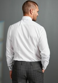 Camicia bianca a maniche lunghe con colletto strutturato, abbinata a pantaloni grigi a quadri con tasche e finitura in tessuto liscia.