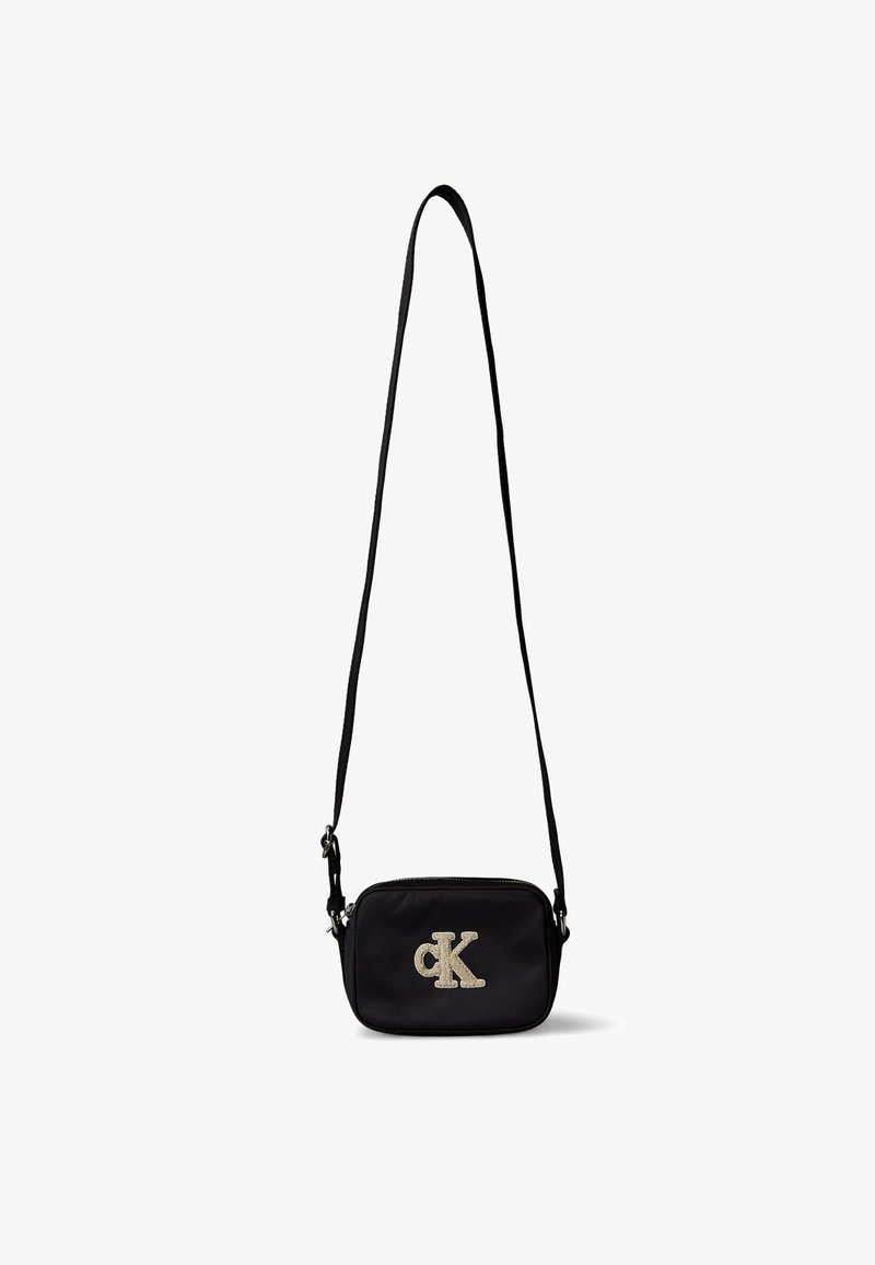 Μικρό μαύρο τσαντάκι ώμου με μακρύ λουράκι και ασημένιο λογότυπο Calvin Klein μπροστά σε λευκό φόντο.