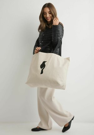 Kobieta w czarnej pikowanej kurtce i beżowych spodniach trzyma dużą beżową torbę na ramię z czarnym sylwetką ptaka na jednolitym tle.