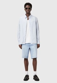 Chemise à rayures bleu clair, débardeur blanc, shorts en denim bleu clair, chaussures noires et lunettes de soleil. Le modèle se tient sur un fond uni.