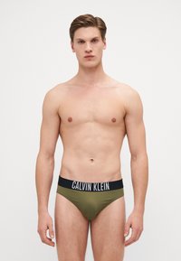 Maslinasto zelena muška gaćice s crnim elastičnim pojasom na kojem se nalazi logo "CALVIN KLEIN". Glatka tekstura i oblikovani dizajn.