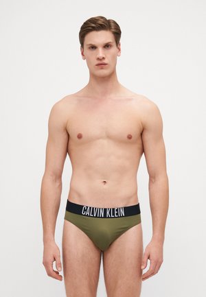 Olivengrønne herretrusser med en sort elastisk livrem med "CALVIN KLEIN" logo. Glat tekstur og tætsiddende design.