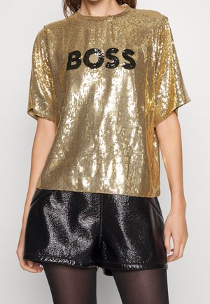 Kobieta w złotej, cekinowej bluzce z krótkim rękawem z napisem „BOSS” oraz błyszczących czarnych szortach z czarnymi rajstopami.