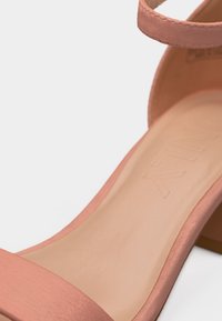 Ροζ σατέν σανδάλι με ανοιχτά δάχτυλα, λουράκι στον αστράγαλο και μεσαίο τακούνι σε σχήμα block, που δείχνει την εσωτερική σόλα και μερικές λεπτομέρειες του τακουνιού.