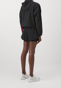 Chaqueta negra corta con capucha, pantalones cortos negros a juego y zapatillas deportivas blancas. Diseño simple, tejido ligero y sin patrones visibles.