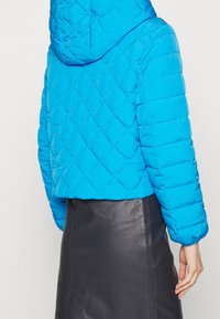 Person som bär en ljusblå vadderad huvrjacka och en svart läderskjol, visad bakifrån mot en enfärgad bakgrund.