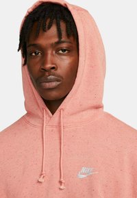 Jasno różowa bluza z kapturem o teksturowanym wykończeniu, z przednią kieszenią, sznurkami oraz małym niebieskim logo Nike na klatce piersiowej.