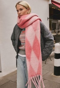 Pink patterned scarf made of soft wool, featuring a diamond design and fringed ends. Worn over a gray quilted jacket and patterned sweater.