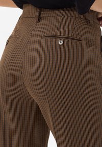 Gros plan sur un pantalon rayé marron et noir avec des poches à l'arrière à boutons, porté par une personne en haut noir.