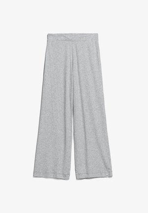 Personne portant un haut gris clair à manches courtes et un pantalon large assorti, debout pieds nus sur un fond uni.